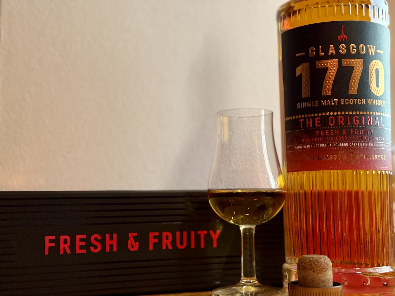 1770 Glasgow The Original Whiskyflasche mit gefülltem Tasting-Glas, aufgenommen seitlich vor hellerer Wand, „Fresh & Fruity“-Box im Vordergrund.