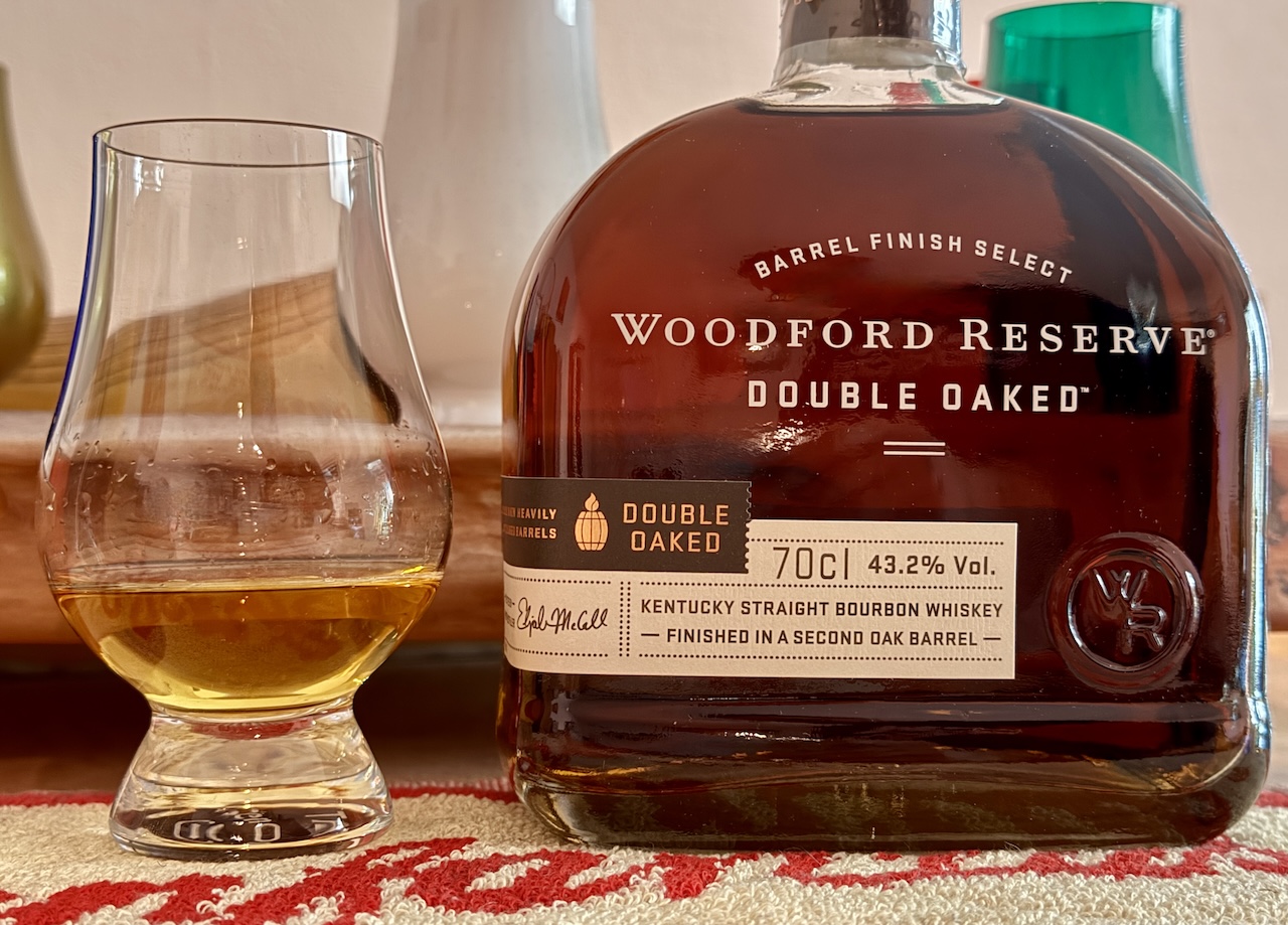 Eine Flasche Woodford Reserve Double Oaked steht neben einem Whiskyglas, beide auf einer roten Unterlage.
